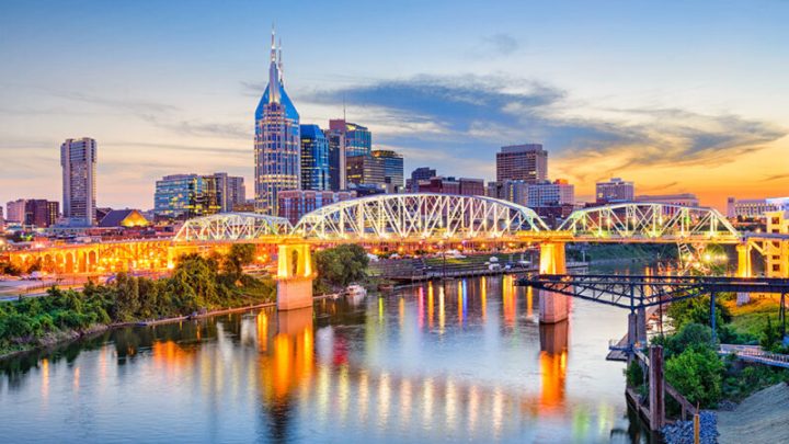 view of Nashville at sunset, showcasing the illuminated John Seigenthaler Pedestrian Bridge spanning over the Cumberland River, with the iconic city skyline, including the AT&T Building, in the background